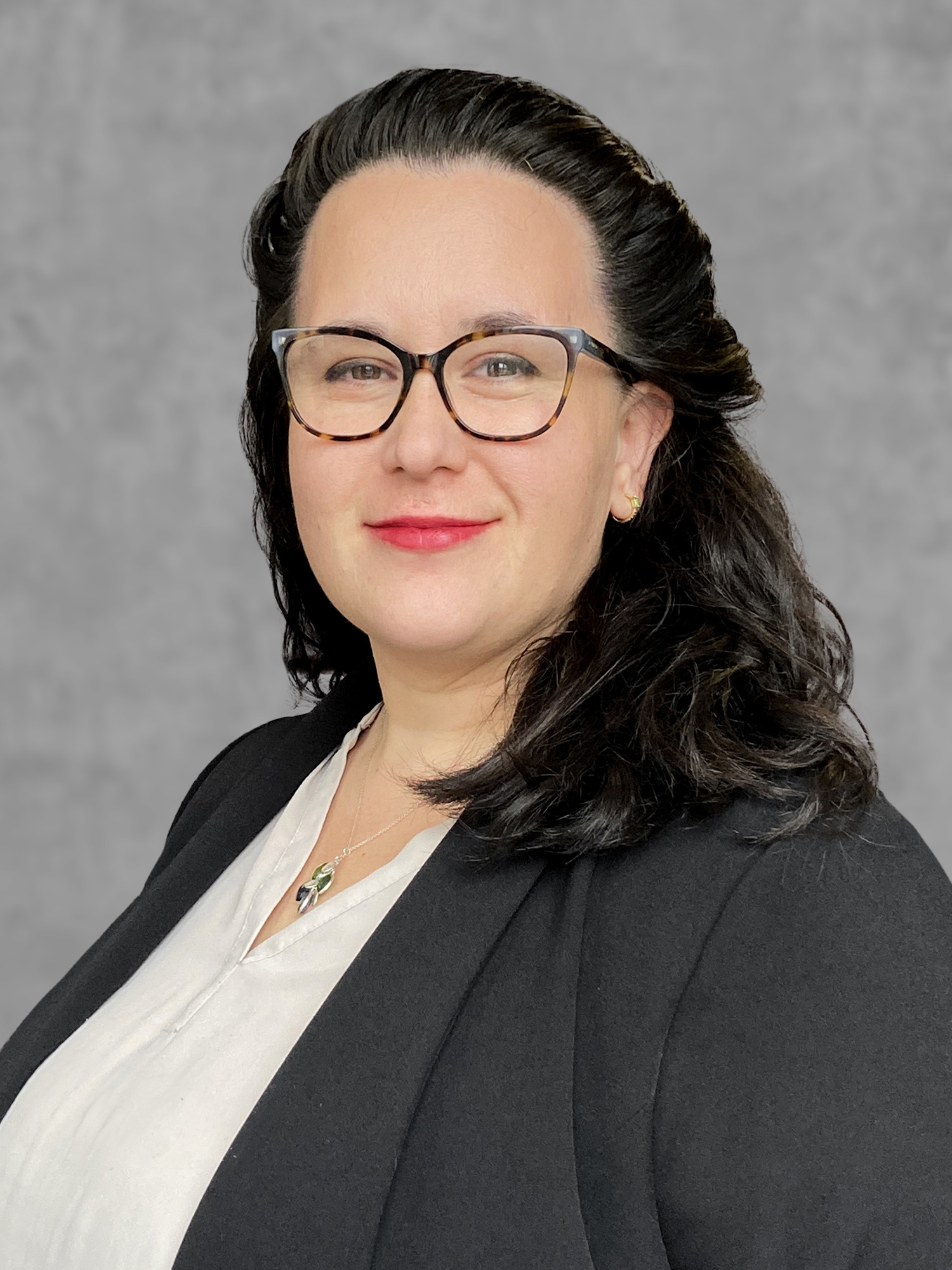 A confident woman with dark hair and glasses, wearing a black blazer and white blouse, smiles against a gray background.