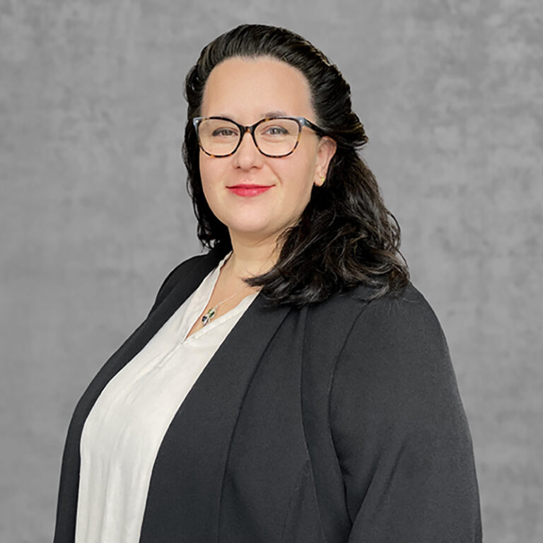 A confident woman with dark hair and glasses, wearing a black blazer and white blouse, smiles against a gray background.