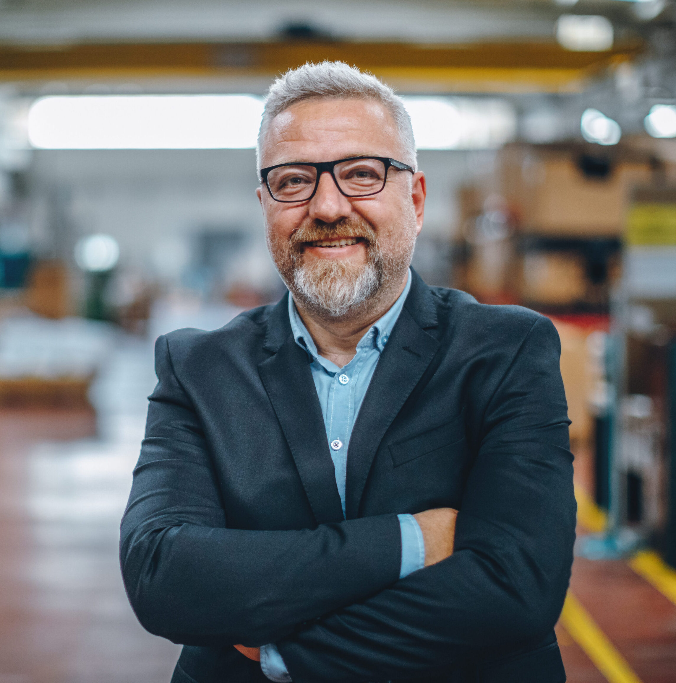Smiling man with glasses and a gray beard, dressed in a suit, stands confidently with arms crossed in a well-lit, spacious warehouse setting.